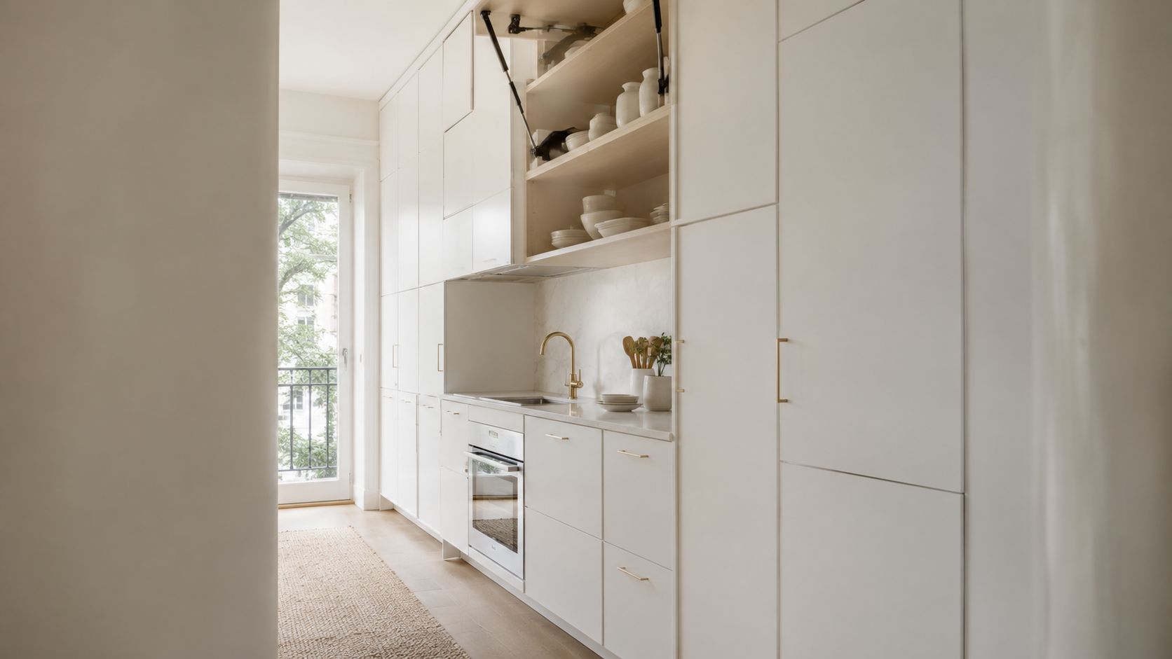 A modern minimalist kitchen with cream cabinetry, brass fixtures, and open shelving in a narrow apartment layout.